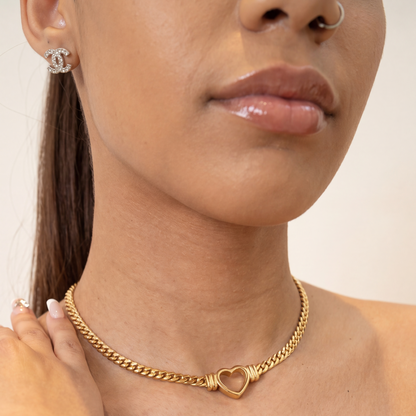 A close-up lifestyle shot of a model wearing a gold chain necklace with a heart-shaped clasp and a matching gold heart bracelet.